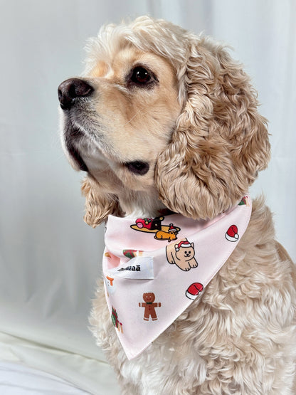 A cream-colored Cocker Spaniel dog wearing a light pink bandana with a holiday print of Santa Claus, gingerbread men, and cookies, set against a plain white background.