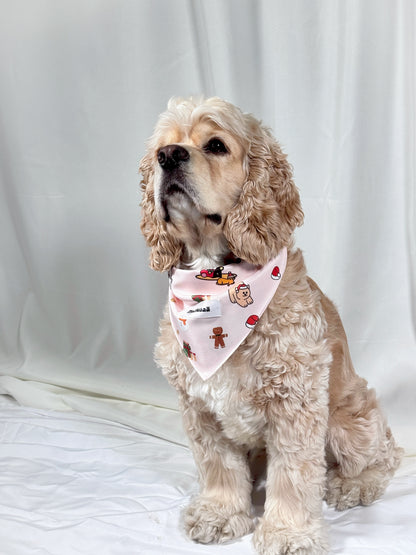 A light cream-colored Cocker Spaniel dog wearing a pink bandana with a gingerbread-themed print, sitting against a white fabric backdrop.