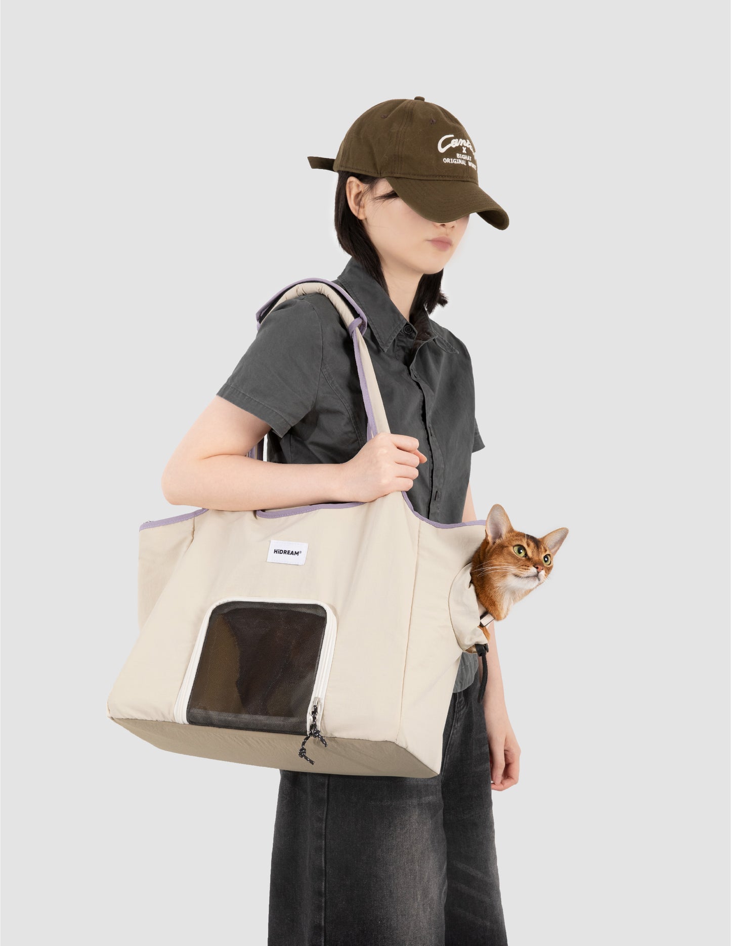 Person holding a beige tote bag with a cat peeking out, wearing a dark cap and gray shirt.