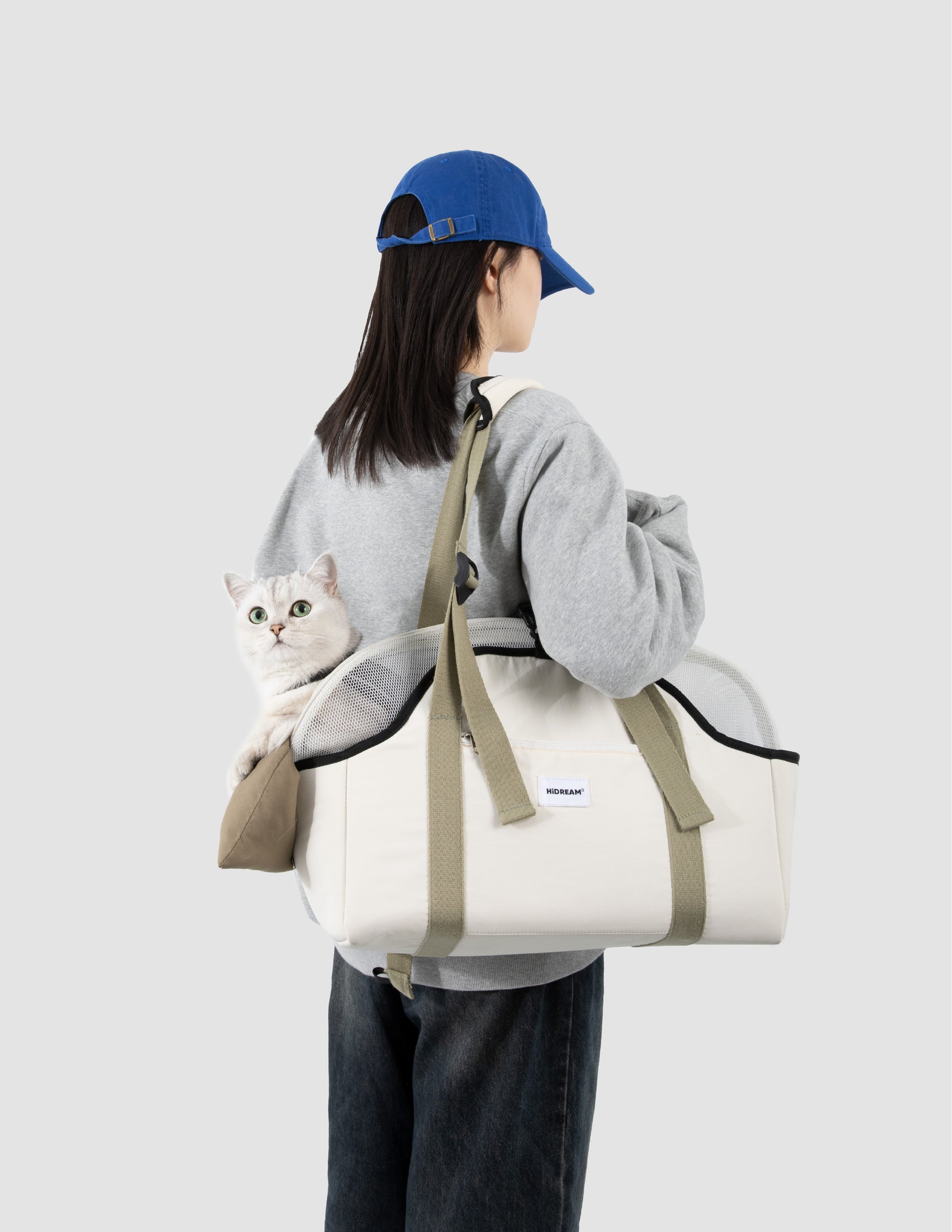 Person carrying a white bag with a gray cat inside, wearing a blue cap and gray hoodie on a light gray background