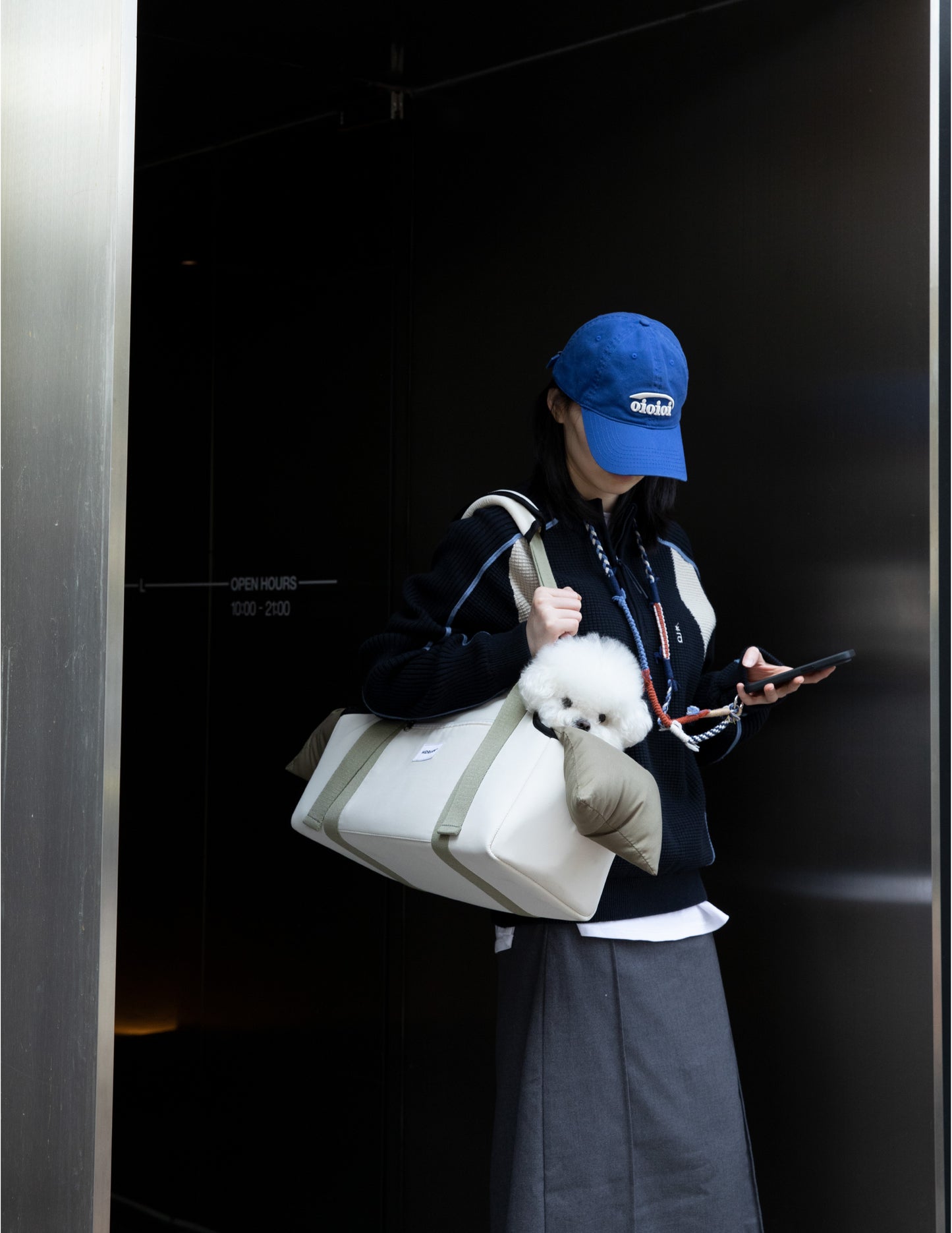 Person holding a small white dog in a bag, wearing a blue cap and dark coat.