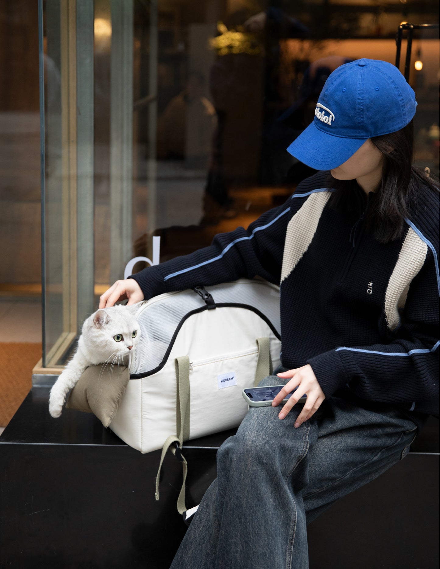 Person holding a large beige tote bag with a pet inside, wearing a cap and headphones, against a gray background.