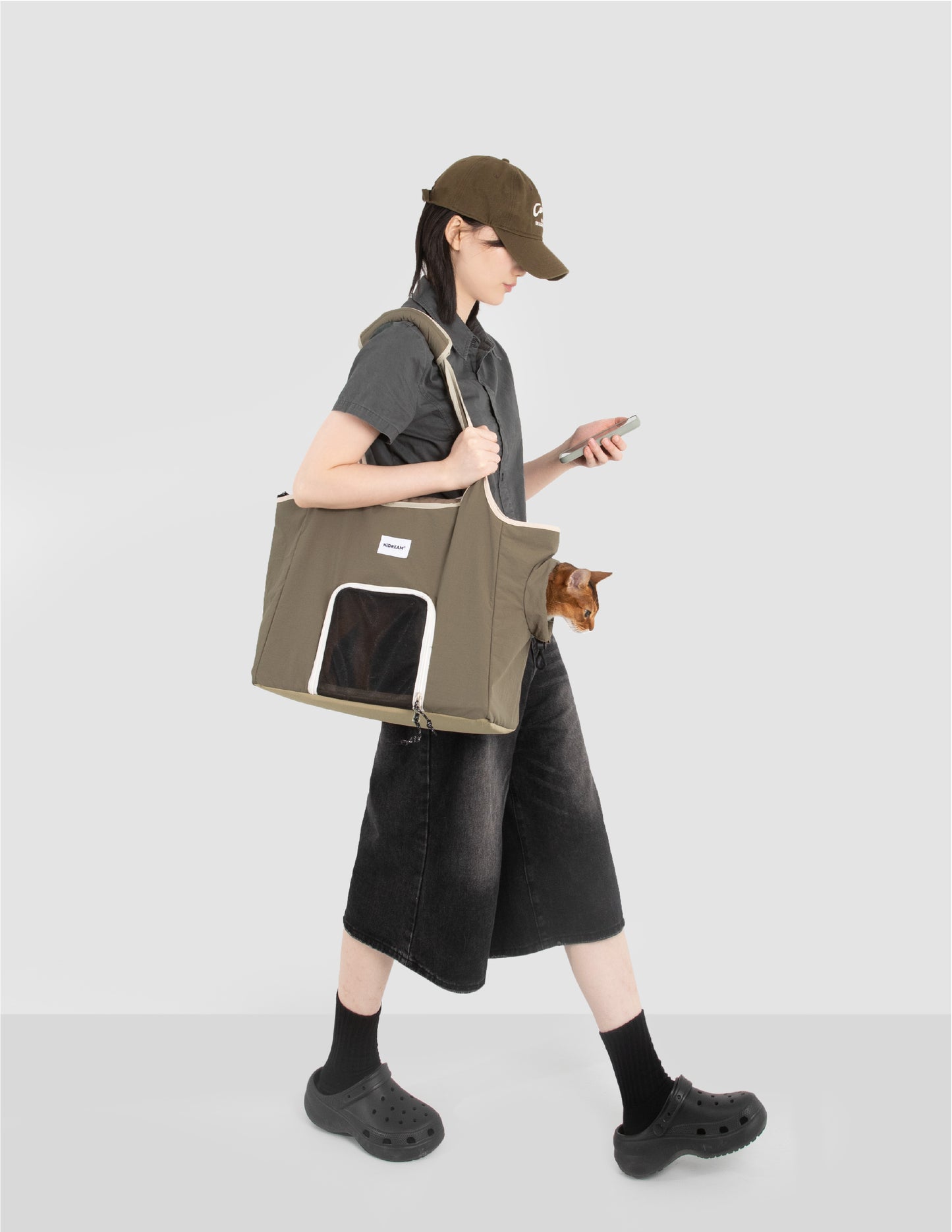 Person carrying an olive green cat carrier tote bag with mesh window while an orange cat peeks out from the side opening.