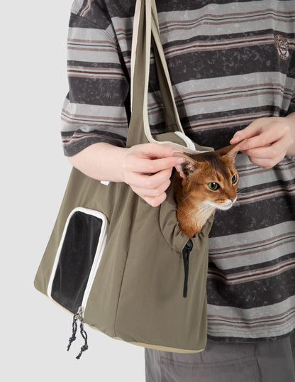 Olive green cat carrier tote bag with side opening and mesh window, holding an orange cat while a person adjusts its ears.