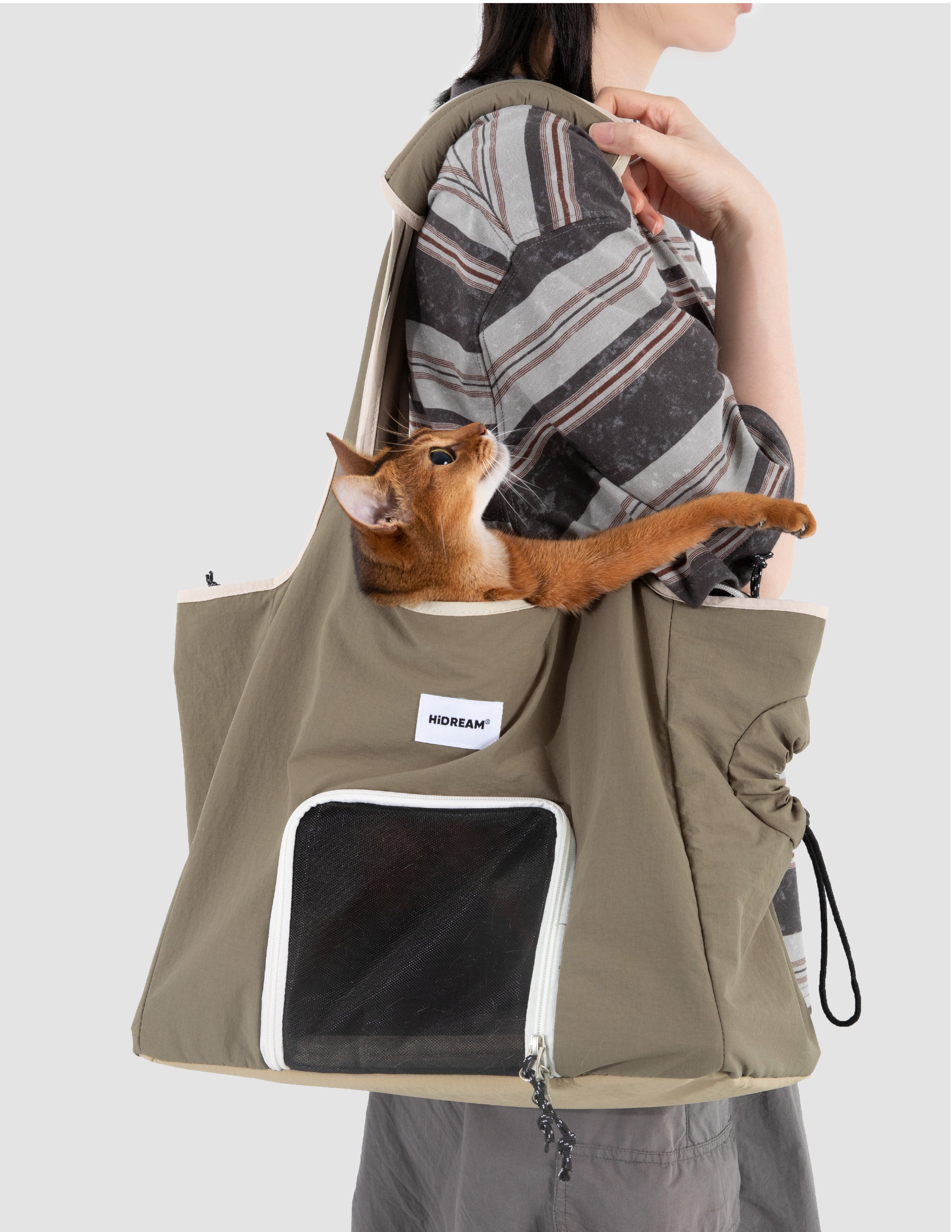 Olive green cat carrier tote bag with mesh front window, carried over the shoulder while an orange cat rests inside and reaches out.