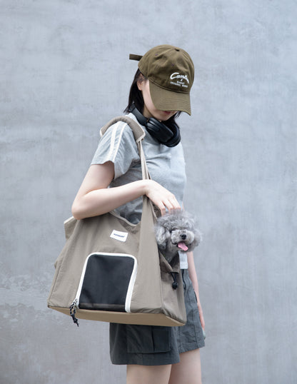 Beige pet carrier tote bag with mesh front window and side opening, carried over the shoulder while a small gray dog looks out.